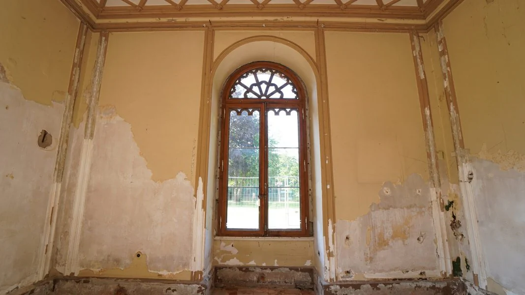 Ein Raum mit einem Fenster und einer Wand, deren Farbe abblättert, vermittelt einen Eindruck von Verfall und Vergänglichkeit.