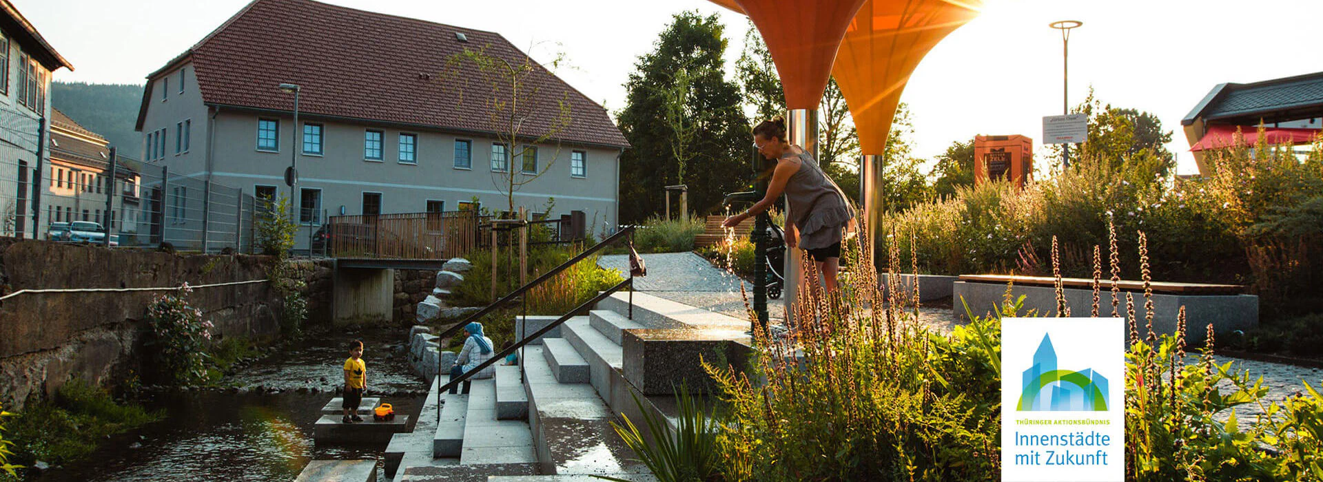Bildbeschreibung / description: Ein Kind mit seinen Eltern spielt bei einer Treppe am Fluss bei Sonnenuntergang in Zella-Mehlis, Logo des Aktionsbündnis Innenstädte unten Rechts