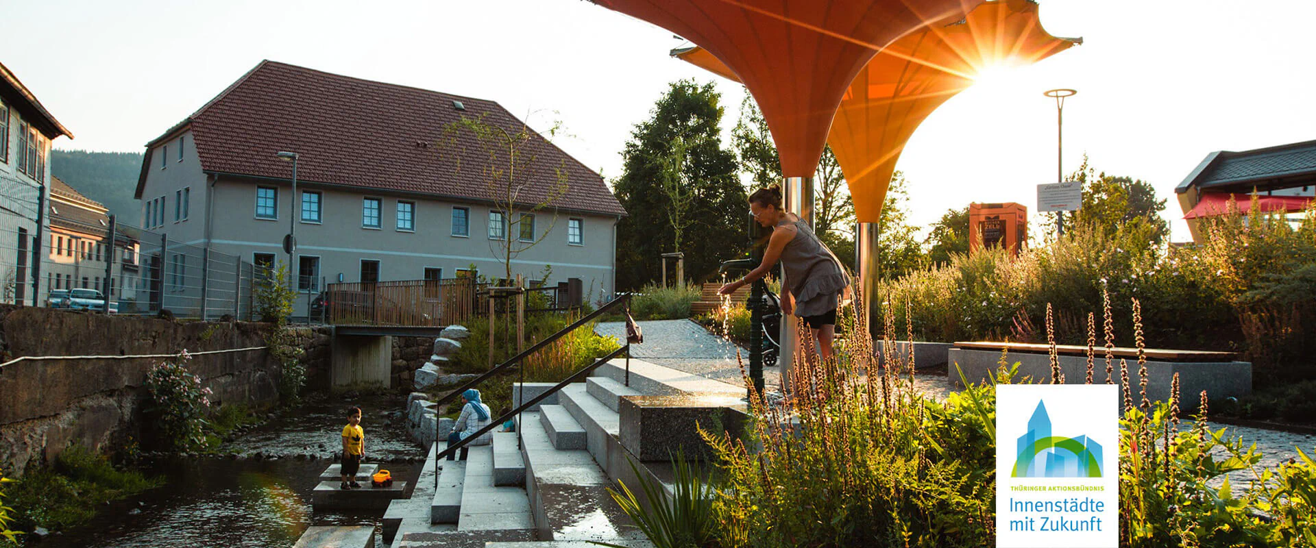 Bildbeschreibung / description: Ein Kind mit seinen Eltern spielt bei einer Treppe am Fluss bei Sonnenuntergang in Zella-Mehlis, Logo des Aktionsbündnis Innenstädte unten Rechts