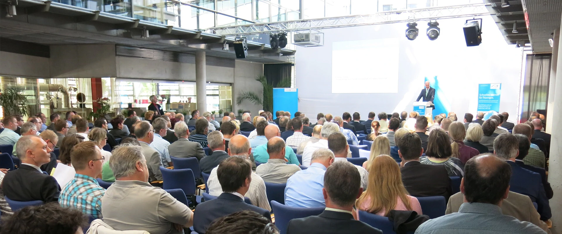 Bildbeschreibung / description: Eine Vielzahl von Menschen ist in einem großen Raum auf Stühlen versammelt und lauscht einem Vortrag