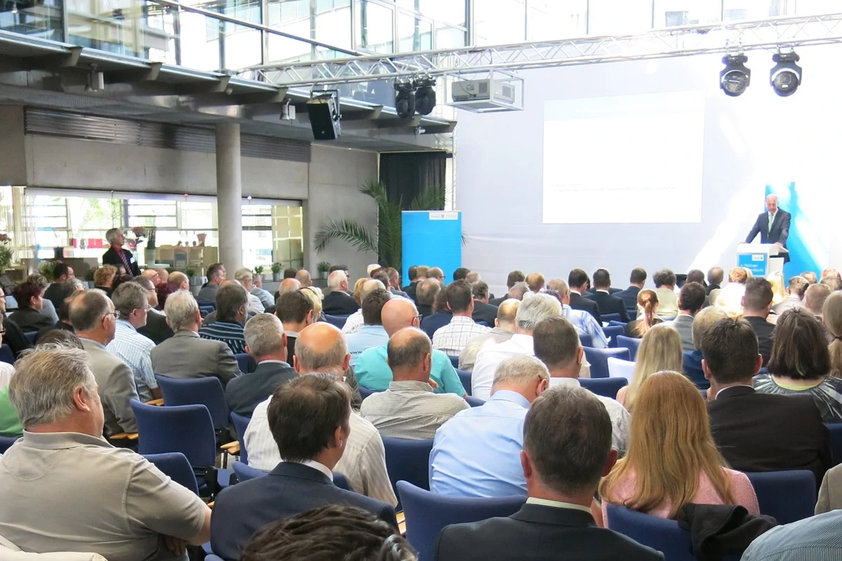 Breitband Bildbeschreibung / description: Eine Vielzahl von Menschen ist in einem großen Raum auf Stühlen versammelt und lauscht einem Vortrag